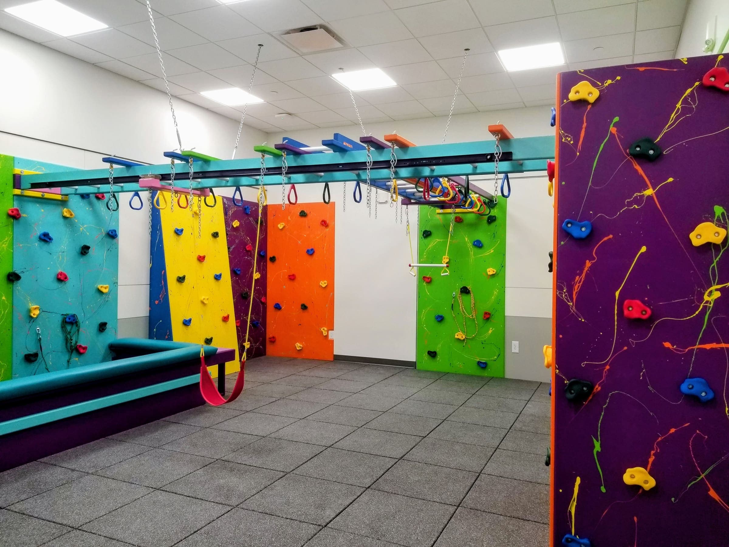Hospital therapy gym with rainbow splatter climbing panels, overhead ring beam, and pink swing