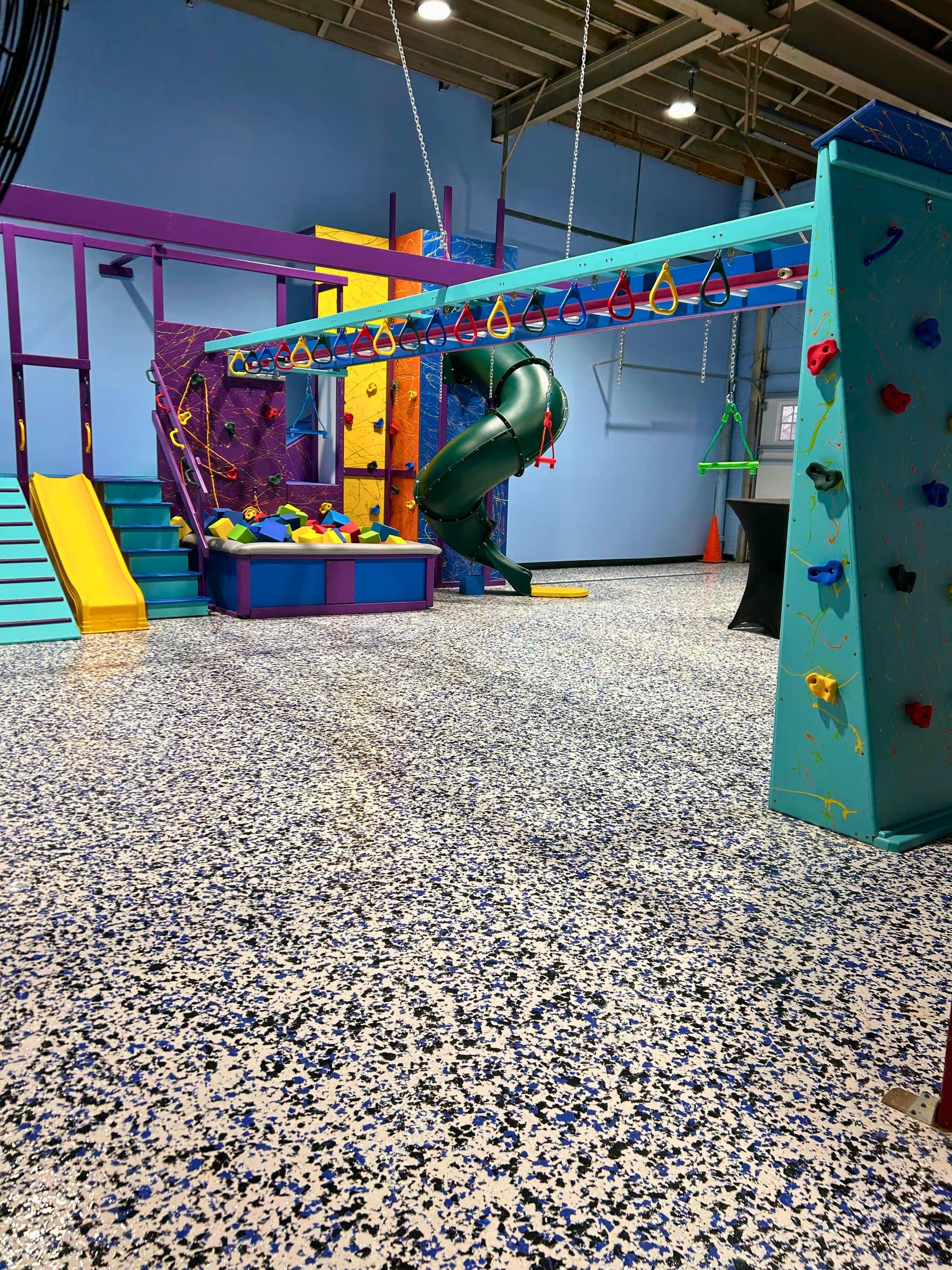 Commercial sensory gym with green tube slide, foam pit, yellow slide, overhead ring beam, and teal climbing wall
