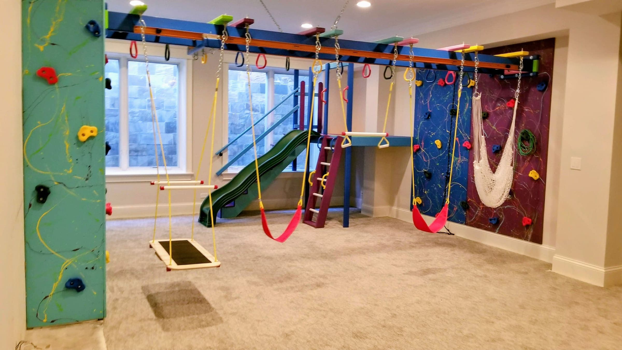 Carpeted basement sensory gym with splatter climbing walls, platform swing, hammock, and green slide